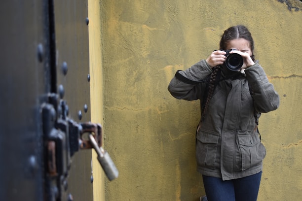 A person holding a camera takes a photograph against a textured yellow wall. They are wearing a green jacket and have long braided hair. To the left, there is a large black metal door with bolts and a padlock, adding an industrial aspect to the scene.