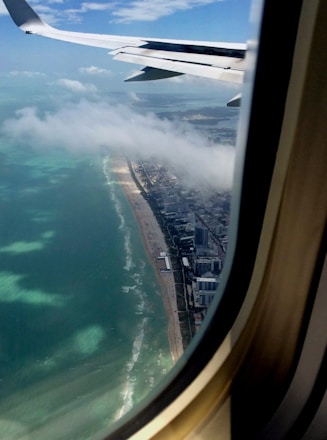 An aerial view from an airplane window showing a coastal cityscape with tall buildings lining the shoreline. The ocean is visible with waves crashing onto the sandy beach. Clouds are floating nearby, and the airplane wing is partially visible.