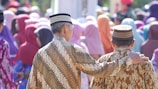 Two people dressed in traditional patterned clothing are standing closely together with one person having their arm around the other. They are surrounded by a crowd of people wearing colorful headscarves and attire, suggesting a social or community gathering.