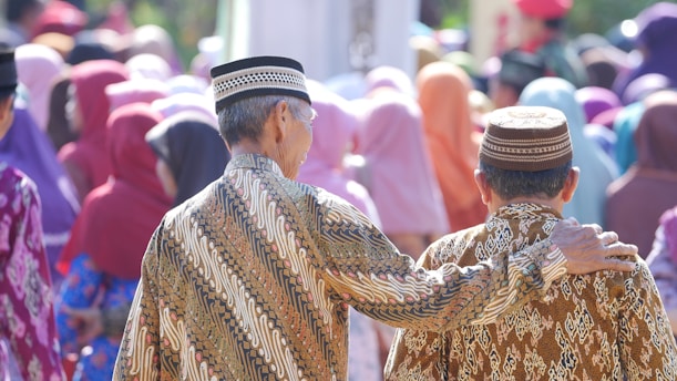 Two people dressed in traditional patterned clothing are standing closely together with one person having their arm around the other. They are surrounded by a crowd of people wearing colorful headscarves and attire, suggesting a social or community gathering.