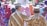 Two people dressed in traditional patterned clothing are standing closely together with one person having their arm around the other. They are surrounded by a crowd of people wearing colorful headscarves and attire, suggesting a social or community gathering.