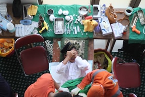 A person is seated in the center with their hands covering their face, surrounded by a variety of medical instruments and supplies on tables. The tables are covered with green cloths, and there are items like syringes, scissors, bandages, and bottles. Two people in orange clothing are interacting with the person in the center. The setting appears to be a temporary medical or clinical setup.