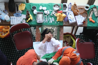 A person is seated in the center with their hands covering their face, surrounded by a variety of medical instruments and supplies on tables. The tables are covered with green cloths, and there are items like syringes, scissors, bandages, and bottles. Two people in orange clothing are interacting with the person in the center. The setting appears to be a temporary medical or clinical setup.
