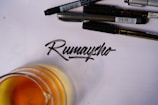 A collection of black pens arranged above a handwritten word in calligraphy on a sheet of paper. An amber-colored liquid in a glass is partially visible at the bottom left of the image.