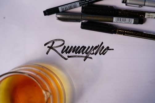 A collection of black pens arranged above a handwritten word in calligraphy on a sheet of paper. An amber-colored liquid in a glass is partially visible at the bottom left of the image.