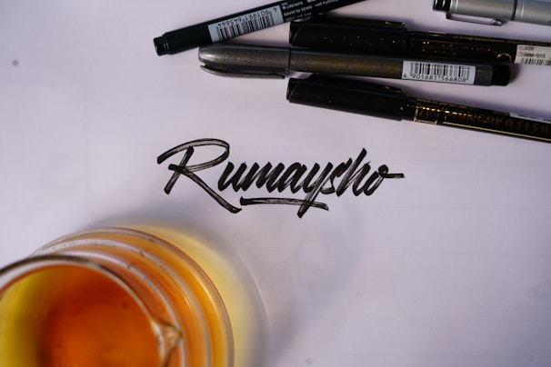 A collection of black pens arranged above a handwritten word in calligraphy on a sheet of paper. An amber-colored liquid in a glass is partially visible at the bottom left of the image.