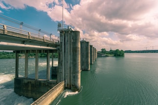 A large concrete dam structure with multiple vertical pillars extends across a flowing river. The water is turbulent, with frothy waves near the structure, and becomes calm further away. In the background, green trees, distant power lines, and cranes are visible under a partly cloudy sky.