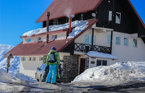 A person wearing colorful ski gear stands next to a snow-covered chalet. The roof is partially covered in snow, with smoke rising from a chimney. The sky is clear and blue, and a car is parked nearby. The person appears to be holding skiing equipment.
