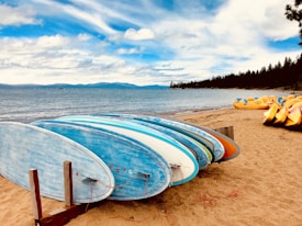 Several surfboards are stacked on a sandy beach with a scenic view of the ocean and distant mountains. The sky is partly cloudy, and additional watercraft are seen in the background.