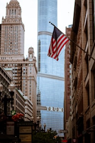 An urban scene featuring a high-rise building with the word 'TRUMP' prominently displayed. The foreground showcases an American flag waving on a pole amidst a street lined with older, ornate architecture. Street signs and building facades are visible along the busy street.