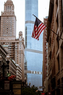 An urban scene featuring a high-rise building with the word 'TRUMP' prominently displayed. The foreground showcases an American flag waving on a pole amidst a street lined with older, ornate architecture. Street signs and building facades are visible along the busy street.