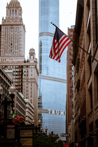 An urban scene featuring a high-rise building with the word 'TRUMP' prominently displayed. The foreground showcases an American flag waving on a pole amidst a street lined with older, ornate architecture. Street signs and building facades are visible along the busy street.