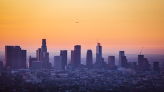 A skyline of tall buildings with a sunset in the background, casting an orange hue across the sky. A small airplane is visible in the air above the cityscape.