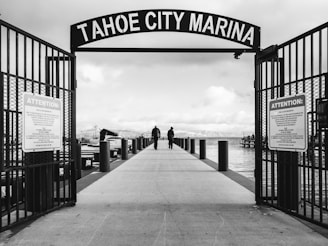 A black and white photograph of a marina entrance with a sign reading 'Tahoe City Marina' at the top. Two people are walking on a wide pier lined with posts, leading out toward a large body of water. The sky is cloudy, and boats are docked to the left. Signs with the word 'Attention' are posted on either side of the entrance gate.