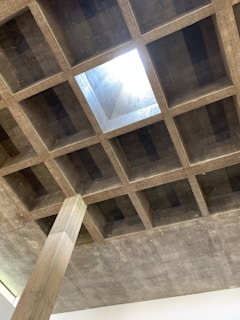 A concrete ceiling features a grid layout with a central square opening allowing light to enter. The structure reveals visible formwork patterns, creating a textured appearance. A vertical concrete column supports the ceiling.