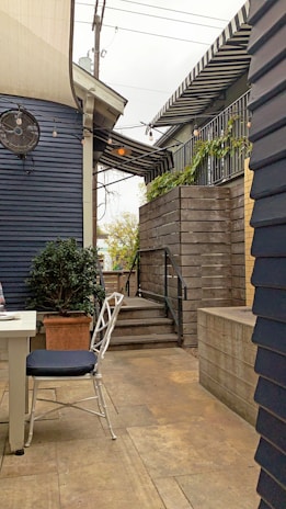 An outdoor patio area with a paved stone floor and wooden walls. A white table and chair with a dark cushion are positioned in the foreground. Stairs lead up to a higher platform adorned with potted plants. Overhead, string lights and a fan are visible under a striped awning.