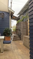 An outdoor patio area with a paved stone floor and wooden walls. A white table and chair with a dark cushion are positioned in the foreground. Stairs lead up to a higher platform adorned with potted plants. Overhead, string lights and a fan are visible under a striped awning.