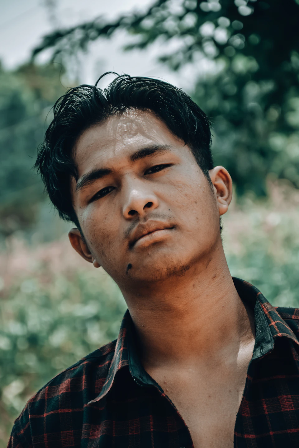 The image shows a young man with dark hair wearing a red and black plaid shirt, standing against a blurred background that appears to be a garden or park setting. The man's gaze is directed off to the side, suggesting he may be lost in thought or observing something outside of the frame.