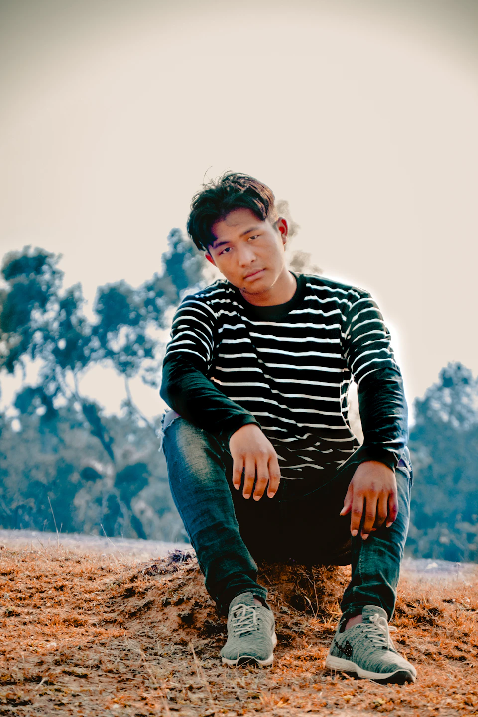 The image shows a young man sitting on the ground, dressed in a black and white striped shirt paired with blue jeans. He is wearing gray sneakers that match his outfit. The background features trees and bushes, creating a natural setting for the scene.