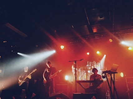 A live music performance featuring a band on stage. Multiple musicians are playing various instruments including guitar and drums. The stage is illuminated with vibrant red and white stage lights creating a dynamic atmosphere.