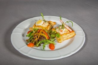 A white plate holds two pieces of puff pastry topped with a creamy salad mixture, garnished with a boiled egg slice and fresh vegetables. The dish is accompanied by a small mixed salad of leafy greens, cherry tomatoes, and bell pepper strips. The setting suggests an elegant presentation on a neutral-colored tablecloth.
