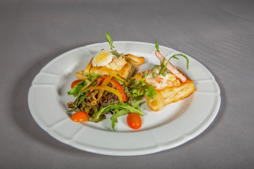 A white plate holds two pieces of puff pastry topped with a creamy salad mixture, garnished with a boiled egg slice and fresh vegetables. The dish is accompanied by a small mixed salad of leafy greens, cherry tomatoes, and bell pepper strips. The setting suggests an elegant presentation on a neutral-colored tablecloth.