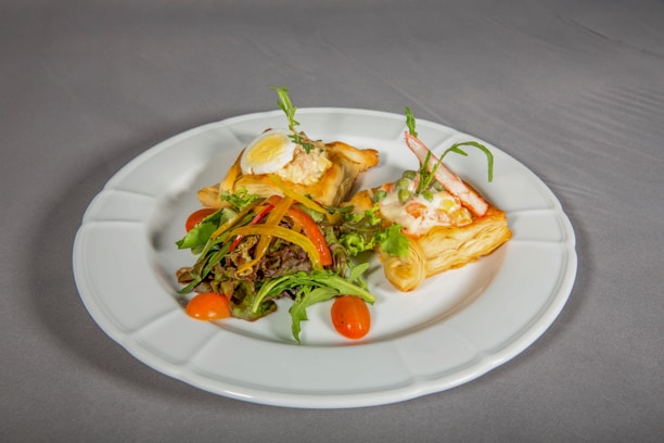 A white plate holds two pieces of puff pastry topped with a creamy salad mixture, garnished with a boiled egg slice and fresh vegetables. The dish is accompanied by a small mixed salad of leafy greens, cherry tomatoes, and bell pepper strips. The setting suggests an elegant presentation on a neutral-colored tablecloth.