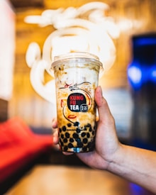 A hand holding a cup of bubble tea from Kung Fu Tea, featuring a creamy and swirled drink with dark tapioca pearls at the bottom. The background is out of focus with warm yellow and blue tones, possibly in a modern café setting.