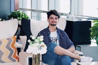 A person sits comfortably on a modern chair in a stylish indoor setting. The individual is dressed in a striped shirt and jeans, with a relaxed and friendly expression. Surrounding the person are various decorative elements, including a vase of white flowers, patterned pillows, and lush green plants. The background features a sleek railing and large glass windows, adding a contemporary feel to the space.