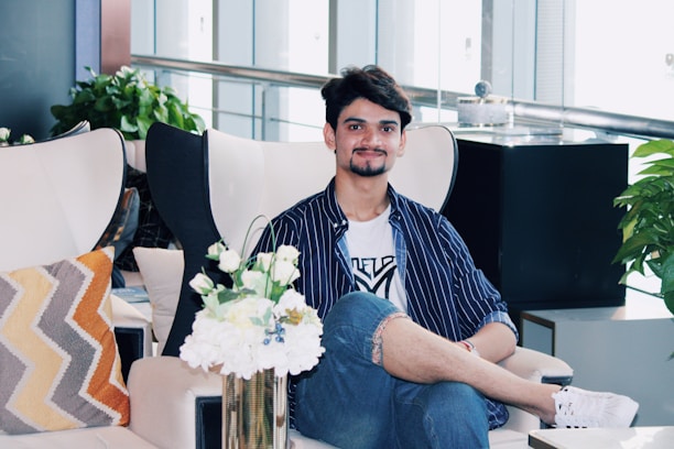 A person sits comfortably on a modern chair in a stylish indoor setting. The individual is dressed in a striped shirt and jeans, with a relaxed and friendly expression. Surrounding the person are various decorative elements, including a vase of white flowers, patterned pillows, and lush green plants. The background features a sleek railing and large glass windows, adding a contemporary feel to the space.
