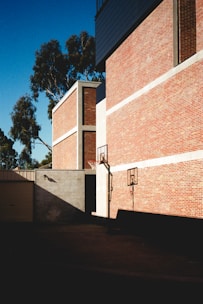 A tall brick building with basketball hoops attached to the side. The structure casts a shadow on the concrete ground in an urban setting. Trees can be seen in the background against a clear blue sky.