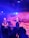A band is performing on stage with three visible musicians. The lead singer holds a microphone, standing in front of a backdrop with the words 'Ciuc' and 'BELUGA'. A guitarist plays on the left, and another musician operates a keyboard in the background. The audience is silhouetted in the foreground under blue and purple lighting.