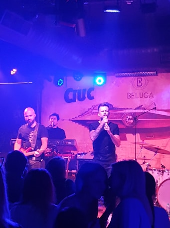 A band is performing on stage with three visible musicians. The lead singer holds a microphone, standing in front of a backdrop with the words 'Ciuc' and 'BELUGA'. A guitarist plays on the left, and another musician operates a keyboard in the background. The audience is silhouetted in the foreground under blue and purple lighting.