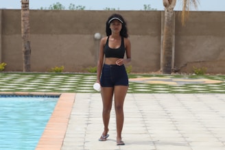 A woman walks confidently alongside a pool, wearing a black sports bra and denim shorts. She holds a small white object and wears a visor on her head. The background features a tiled pool area, grass, and a concrete wall with palm trees.
