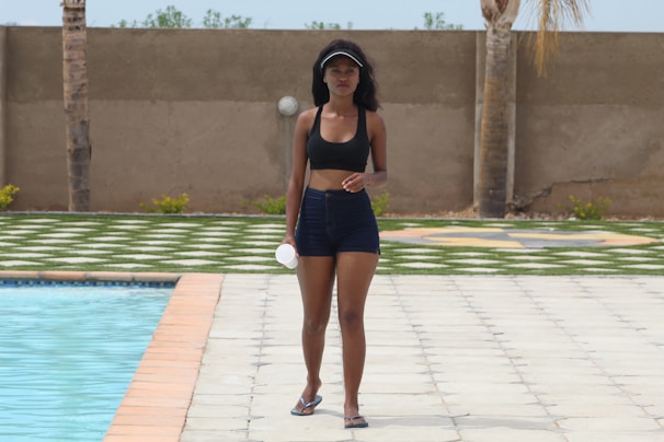 A woman walks confidently alongside a pool, wearing a black sports bra and denim shorts. She holds a small white object and wears a visor on her head. The background features a tiled pool area, grass, and a concrete wall with palm trees.
