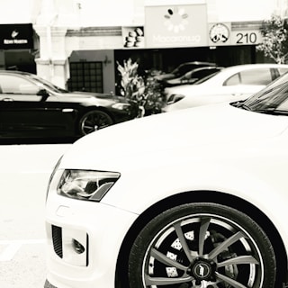 A black and white image featuring the front end of a white car prominently in the foreground, showcasing the vehicle's headlight, grille, and wheel. In the background, there are two other cars, a black one and a white one, parked along a street. Various storefront signs are visible, including one with colorful macarons depicted in grayscale.