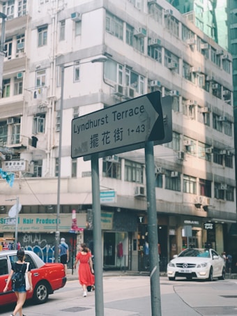 A city street scene in front of an older residential building, with a street sign indicating Lyndhurst Terrace in both English and Chinese. Pedestrians are walking, including a person in a vibrant red dress and a woman carrying a shoulder bag. Two cars are visible on the street, one of which is a red taxi, the other a white Mercedes. The area has a few shops such as a hair products store, and the facade of the buildings shows signs of wear and age.