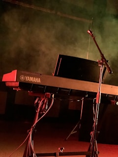 A Yamaha keyboard on a stand illuminated by dramatic stage lighting, with a microphone stand positioned nearby. The background is filled with smoky haze, suggesting a performance or rehearsal setting.