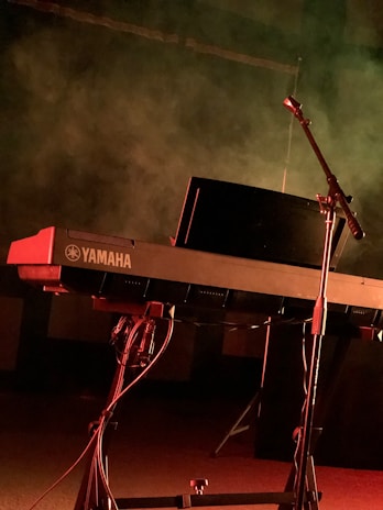 A Yamaha keyboard on a stand illuminated by dramatic stage lighting, with a microphone stand positioned nearby. The background is filled with smoky haze, suggesting a performance or rehearsal setting.