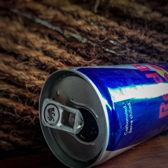 A close-up view of a metallic can laid on its side against a textured background. The can is mostly blue with some writing visible. The can's opening tab is visible, and the background seems to be made of brown, fibrous material.