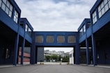 A modern industrial or commercial building with a blue facade, featuring an arched passageway that connects two structures. The architecture is characterized by large windows and minimalistic design. The background includes a residential area with trees under a cloudy sky.