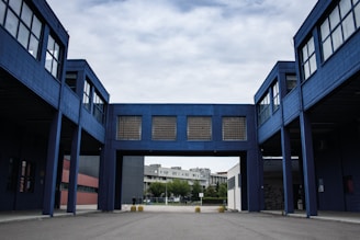 A modern industrial or commercial building with a blue facade, featuring an arched passageway that connects two structures. The architecture is characterized by large windows and minimalistic design. The background includes a residential area with trees under a cloudy sky.