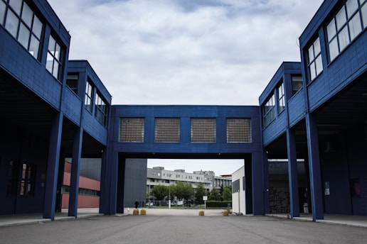 A modern industrial or commercial building with a blue facade, featuring an arched passageway that connects two structures. The architecture is characterized by large windows and minimalistic design. The background includes a residential area with trees under a cloudy sky.