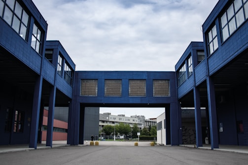 A modern industrial or commercial building with a blue facade, featuring an arched passageway that connects two structures. The architecture is characterized by large windows and minimalistic design. The background includes a residential area with trees under a cloudy sky.