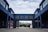 A modern industrial or commercial building with a blue facade, featuring an arched passageway that connects two structures. The architecture is characterized by large windows and minimalistic design. The background includes a residential area with trees under a cloudy sky.