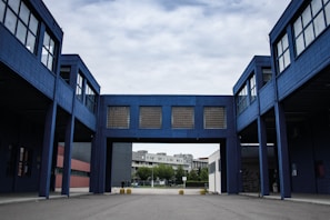 A modern industrial or commercial building with a blue facade, featuring an arched passageway that connects two structures. The architecture is characterized by large windows and minimalistic design. The background includes a residential area with trees under a cloudy sky.