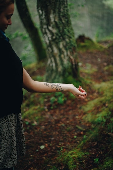 A person is standing in a forest, extending their arm which is adorned with a floral tattoo. The surrounding environment is lush with moss, trees, and earthy tones.