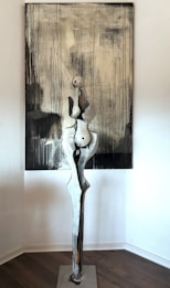 A tall, abstract sculpture is standing in front of a large painting. The sculpture features elongated, curved shapes resembling a humanoid form, with a rustic and textured appearance. The backdrop consists of a monochrome painting with vertical black drips against a gray and white background, creating an impression of rain or movement.