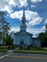 A white church with a tall steeple is set against a vibrant blue sky filled with scattered clouds. The building features classical architectural elements, including columns and a triangular pediment. Surrounding the church are lush green trees and a well-maintained lawn with some flowers. A sign in front of the church provides information about upcoming events.