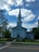 A white church with a tall steeple is set against a vibrant blue sky filled with scattered clouds. The building features classical architectural elements, including columns and a triangular pediment. Surrounding the church are lush green trees and a well-maintained lawn with some flowers. A sign in front of the church provides information about upcoming events.